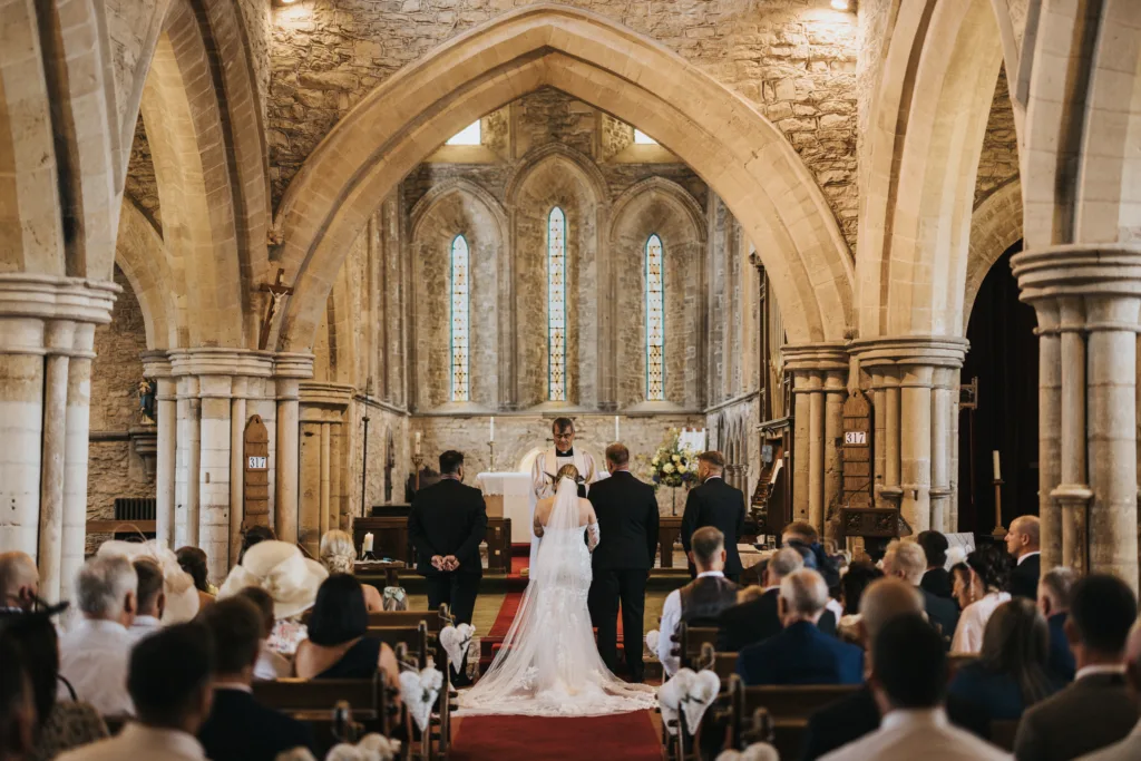 A bride and groom stand at the altar in a historic stone church at Elsham Hall, flanked by attendants as the wedding photographer captures every moment. Warm light streams through stained-glass windows, highlighting the arched architecture. © Aimee Lince Photography, Lincolnshire photographer