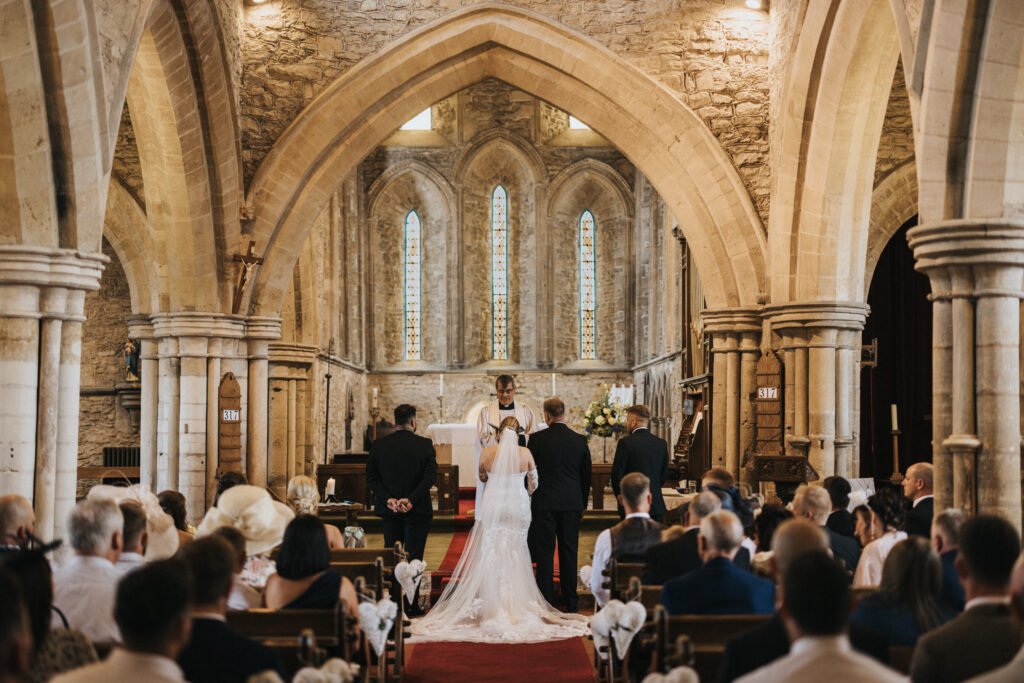 A bride and groom stand at the altar in a historic stone church at Elsham Hall, flanked by attendants as the wedding photographer captures every moment. Warm light streams through stained-glass windows, highlighting the arched architecture. © Aimee Lince Photography, Lincolnshire photographer