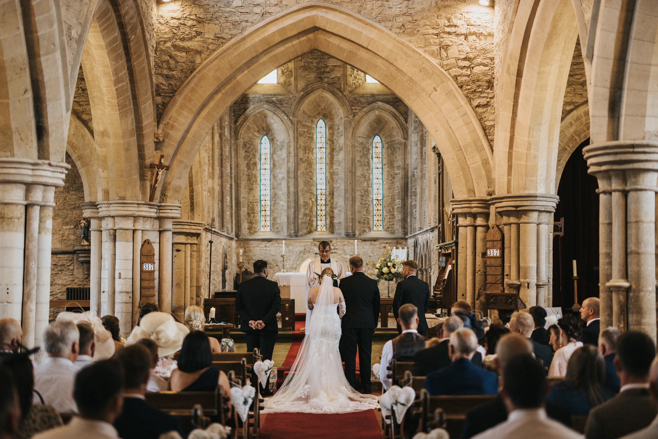 A bride and groom stand at the altar in a historic stone church at Elsham Hall, flanked by attendants as the wedding photographer captures every moment. Warm light streams through stained-glass windows, highlighting the arched architecture. © Aimee Lince Photography, Lincolnshire photographer