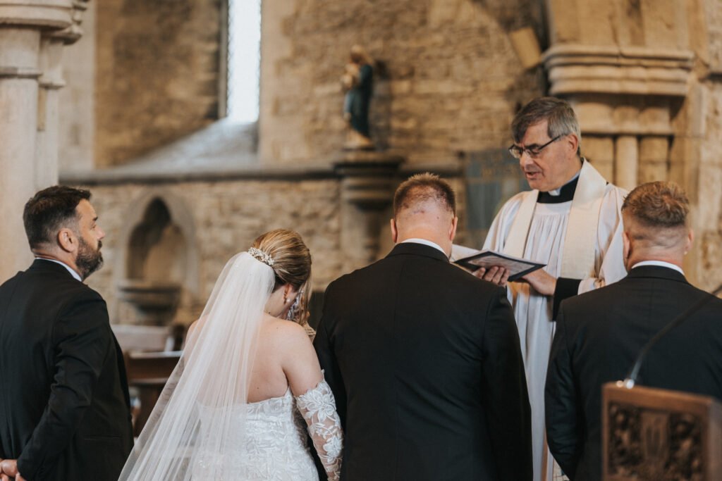 A bride and groom stand before a priest in a stone church at Elsham Hall during their wedding ceremony. A wedding photographer captures the moment as the bride in a white dress and veil listens, while the groom and groomsmen wear black suits. © Aimee Lince Photography, Lincolnshire photographer