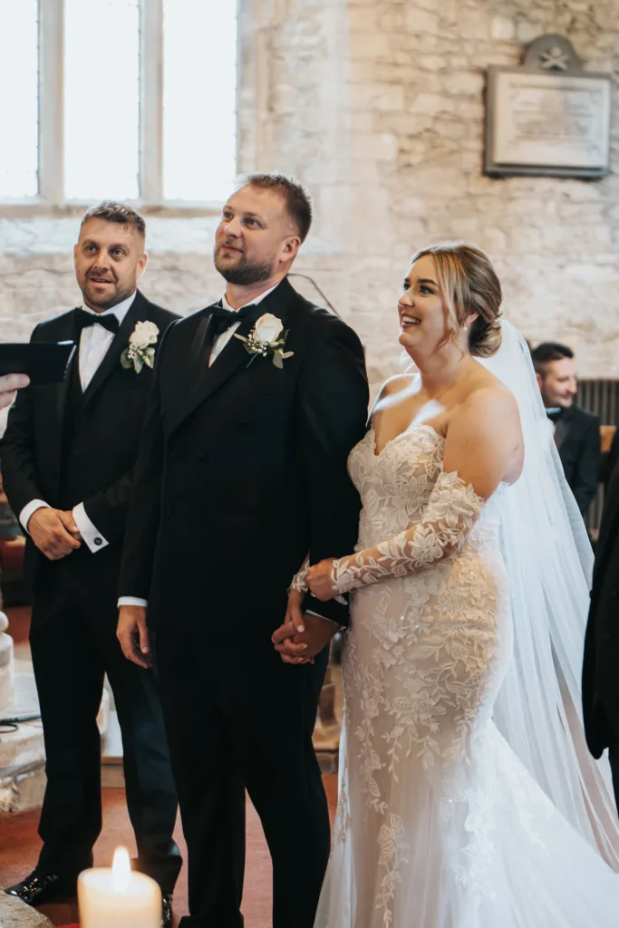A bride in a lace, off-the-shoulder wedding dress and veil stands holding hands with the groom, captured by a wedding photographer as they smile during their ceremony inside a stone church at Elsham Hall, with an usher in the background. © Aimee Lince Photography, Lincolnshire photographer