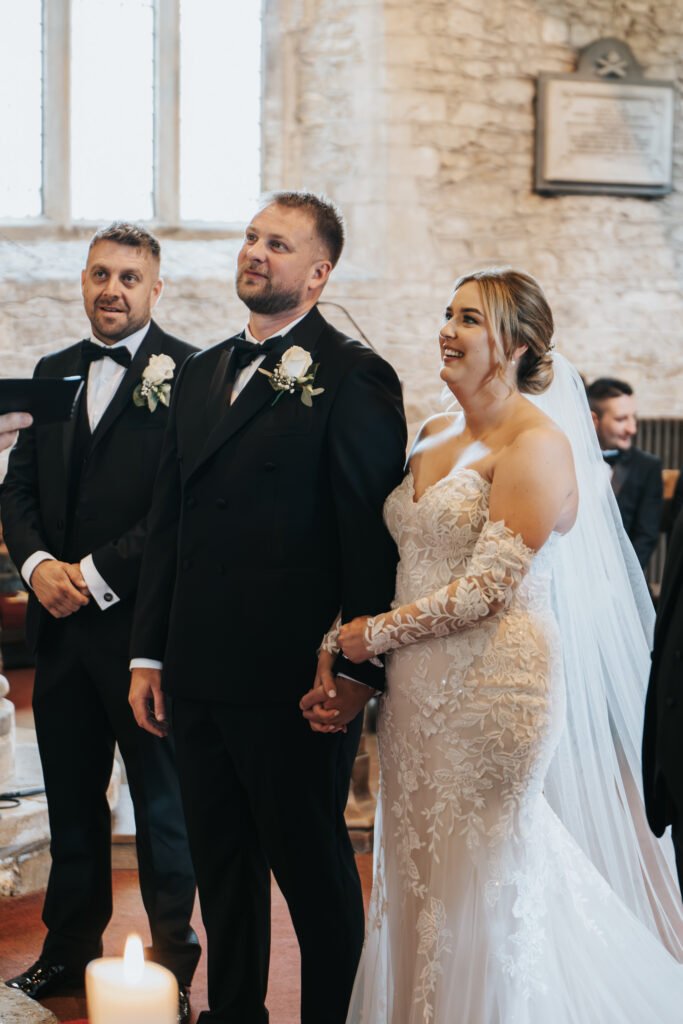 A bride in a lace, off-the-shoulder wedding dress and veil stands holding hands with the groom, captured by a wedding photographer as they smile during their ceremony inside a stone church at Elsham Hall, with an usher in the background. © Aimee Lince Photography, Lincolnshire photographer