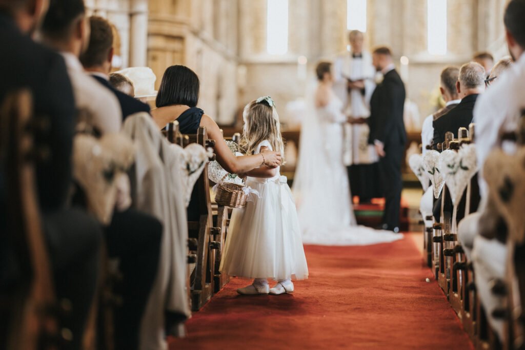 A young flower girl in a white dress stands in the aisle of Elsham Hall’s church, holding a basket. Guests sit on decorated chairs, and a woman gently holds her arm. A wedding photographer captures the bride and groom at the altar, softly out of focus. © Aimee Lince Photography, Lincolnshire photographer