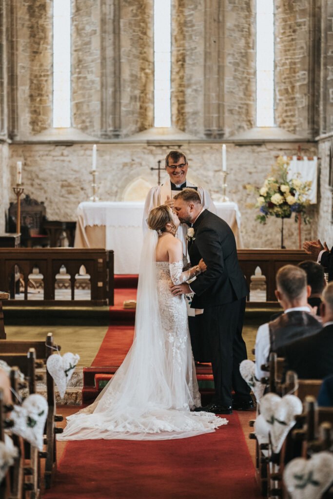 A wedding photographer captures a bride and groom sharing a kiss at the altar in a sunlit church, with guests watching from pews. The bride's lace gown and veil complement the groom’s black suit as a smiling vicar looks on. © Aimee Lince Photography, Lincolnshire photographer