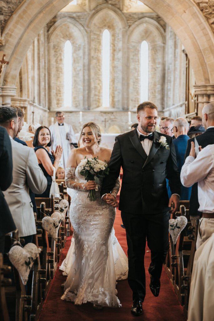 A bride in a white lace gown and groom in a black tuxedo walk hand in hand down the aisle of a stone church at Elsham Hall, smiling as guests applaud, whilst a wedding photographer captures the heart-shaped details and warm natural light. © Aimee Lince Photography, Lincolnshire photographer