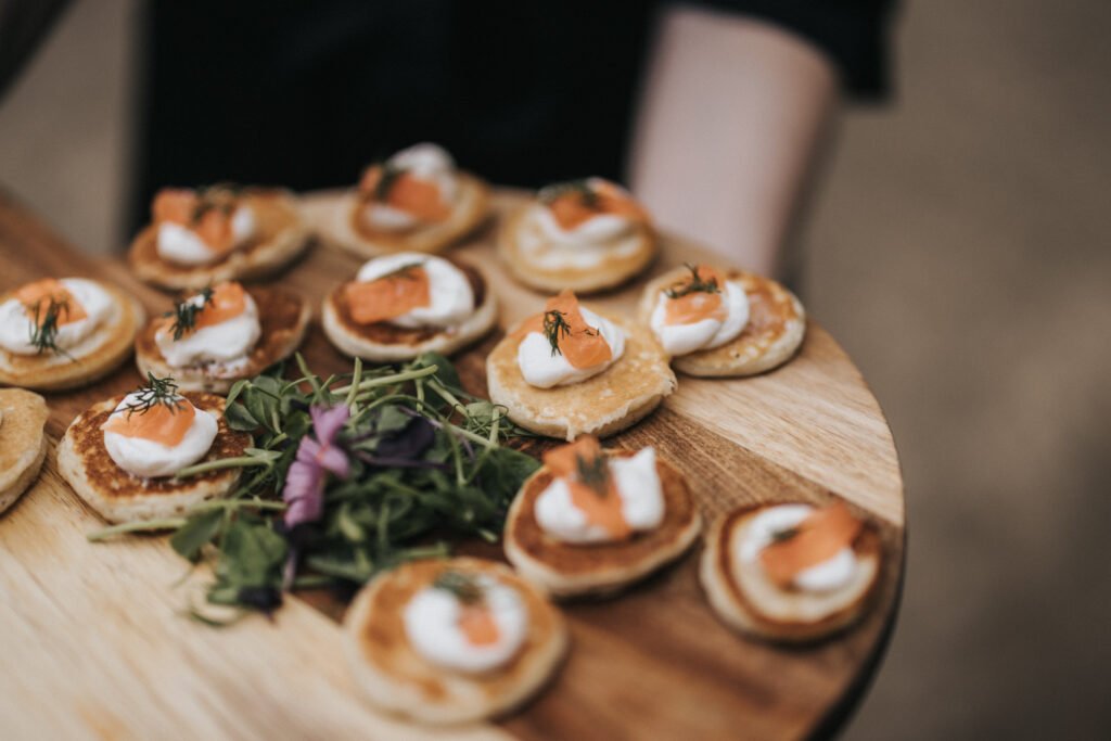 A wooden platter holds several small blinis topped with soured cream, smoked salmon, and dill, arranged neatly around a garnish of fresh greens and edible flowers—perfect for an Elsham Hall wedding photographer to capture. The background is blurred for focus. © Aimee Lince Photography, Lincolnshire photographer