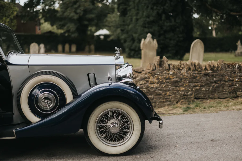 A vintage car with whitewall tyres and chrome details is parked on a paved road. Behind it, an old stone wall and weathered gravestones evoke a calm, historic scene—ideal inspiration for a wedding photographer at Elsham Hall. © Aimee Lince Photography, Lincolnshire photographer