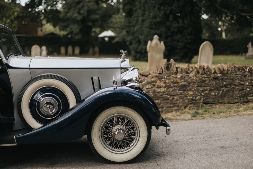 A vintage car with whitewall tyres and chrome details is parked on a paved road. Behind it, an old stone wall and weathered gravestones evoke a calm, historic scene—ideal inspiration for a wedding photographer at Elsham Hall. © Aimee Lince Photography, Lincolnshire photographer