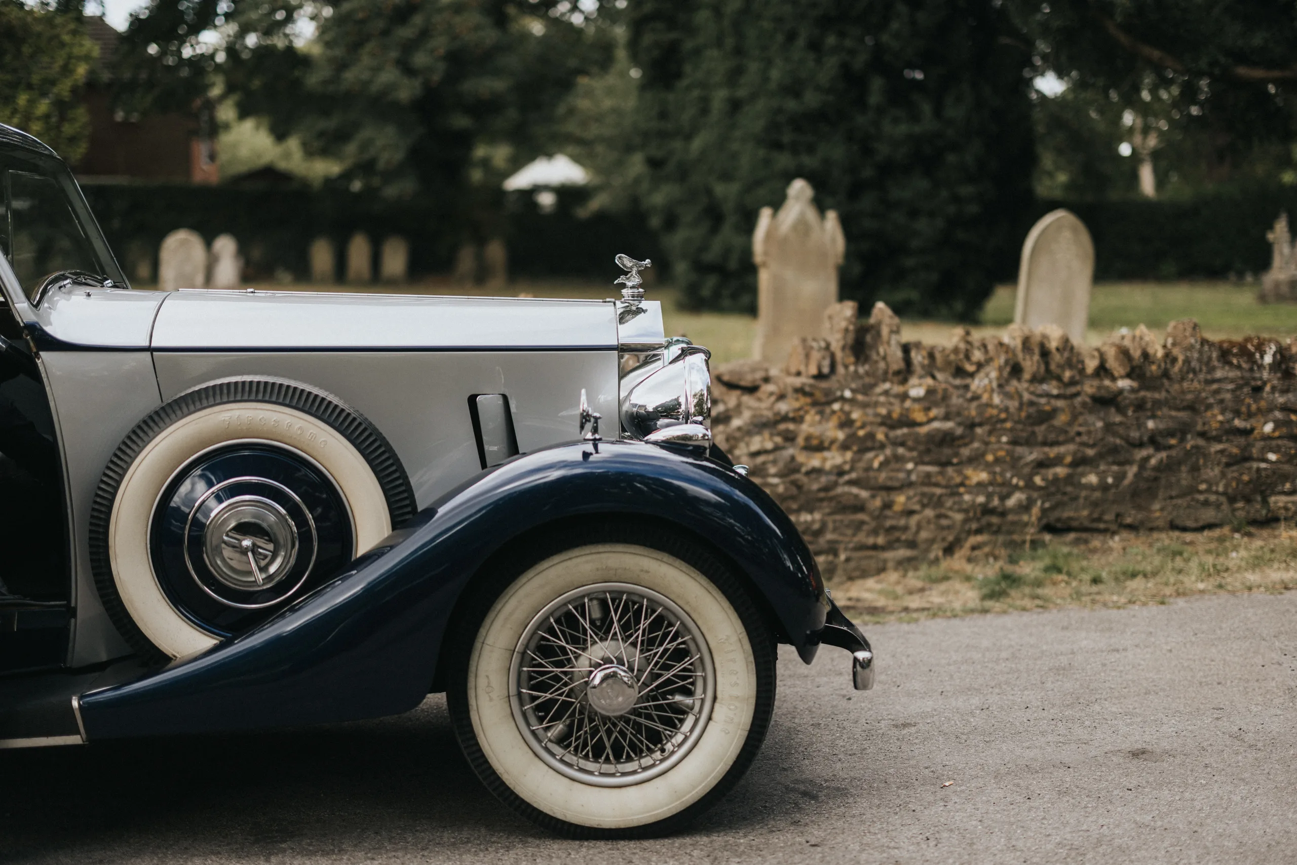 A vintage car with whitewall tyres and chrome details is parked on a paved road. Behind it, an old stone wall and weathered gravestones evoke a calm, historic scene—ideal inspiration for a wedding photographer at Elsham Hall. © Aimee Lince Photography, Lincolnshire photographer