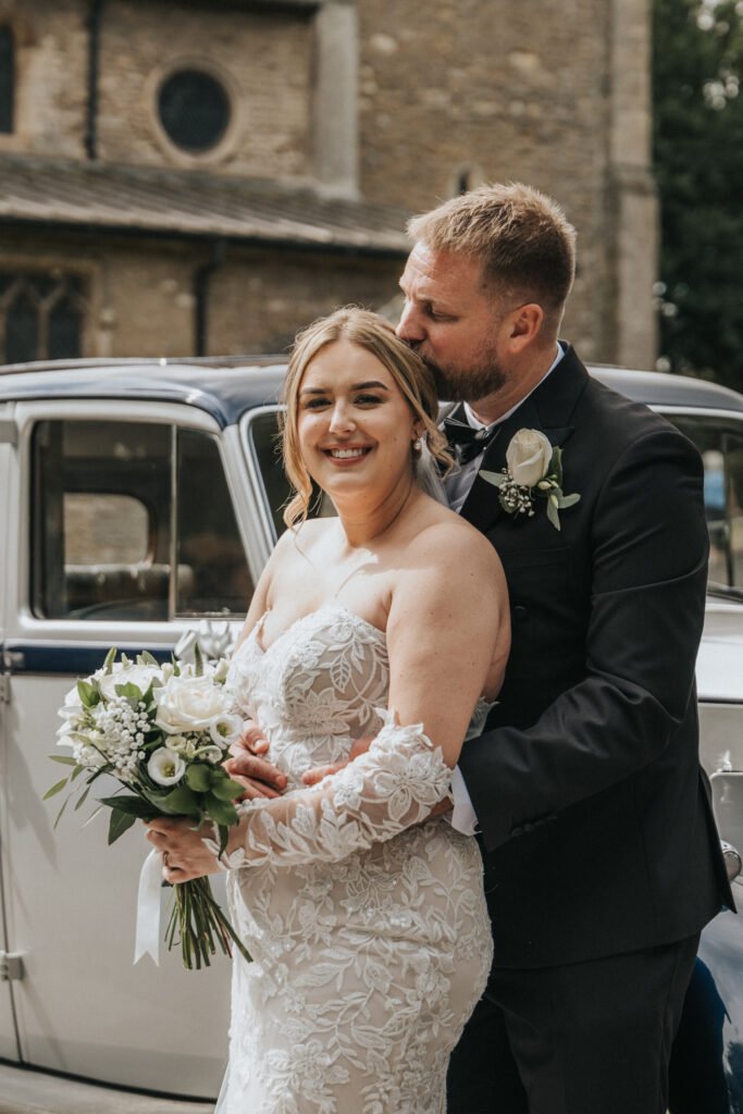 A bride in a white lace wedding dress smiles whilst holding a bouquet of white flowers. A groom in a black suit stands behind her, embracing and kissing her head. Captured by a wedding photographer at Elsham Hall, with a classic car and stone building behind them. © Aimee Lince Photography, Lincolnshire photographer
