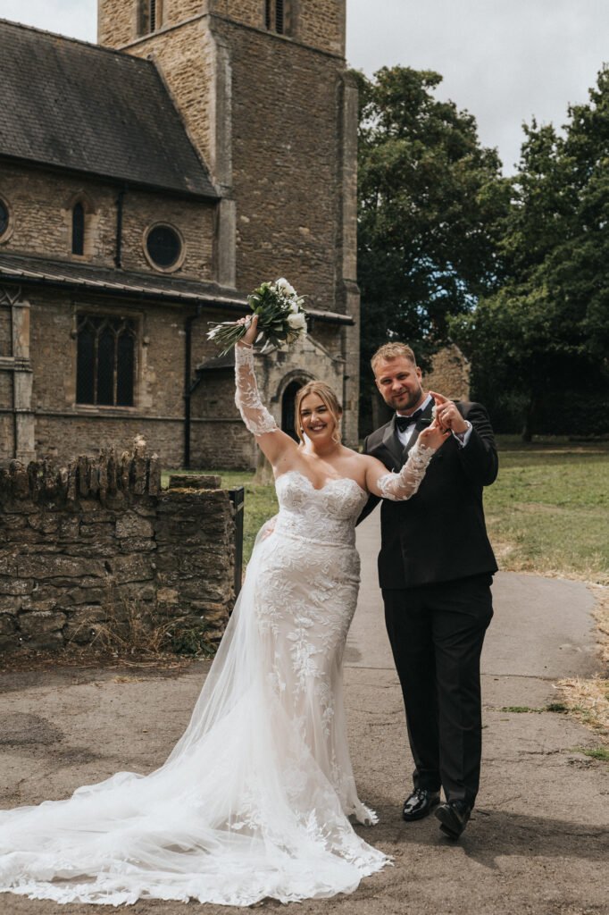 A bride in a white lace gown and a groom in a black tuxedo stand outside Elsham Hall’s stone church. The smiling bride holds her bouquet aloft, whilst the groom gives a thumbs up—perfect moments for any wedding photographer to capture. © Aimee Lince Photography, Lincolnshire photographer