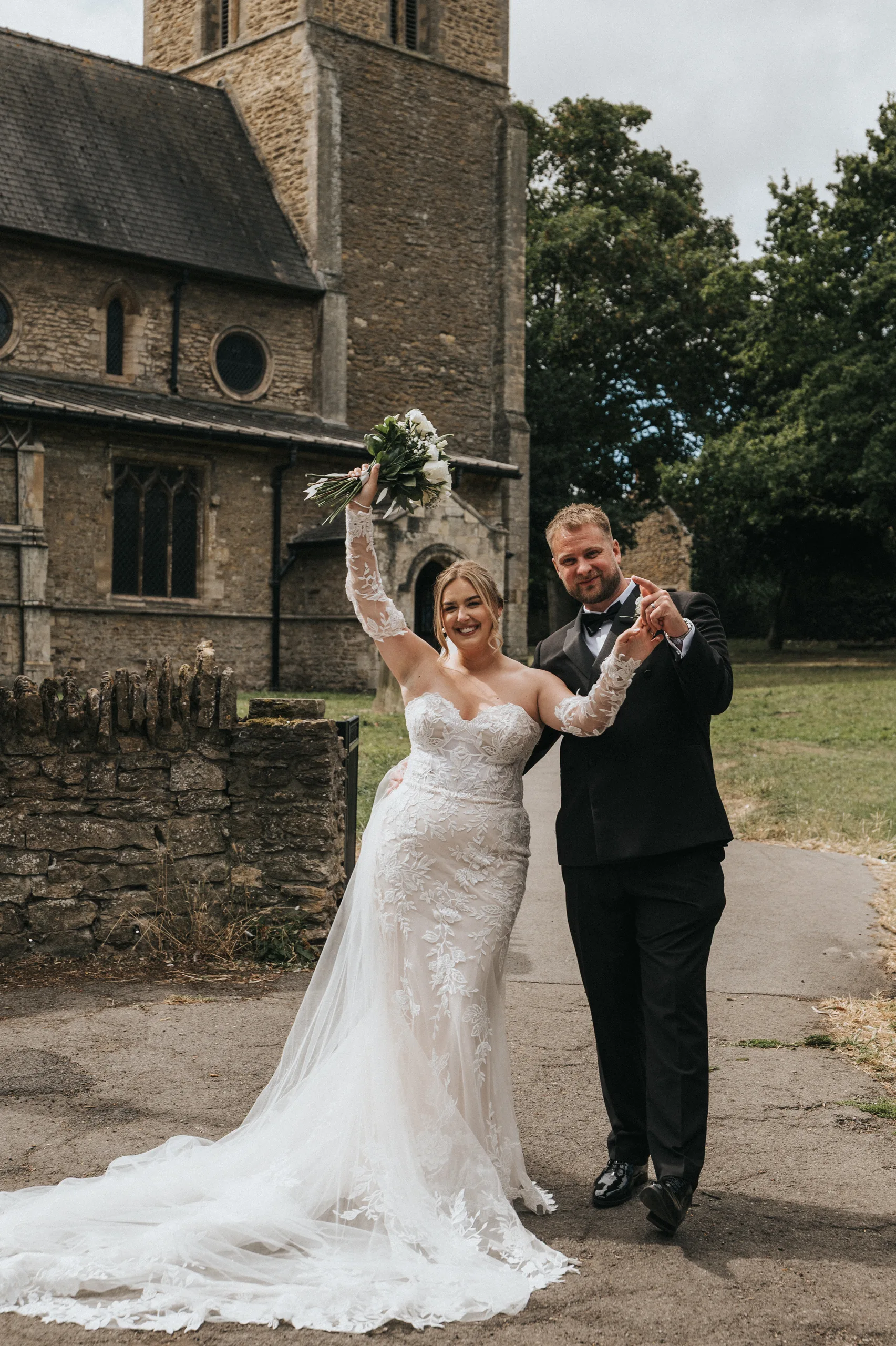 A bride in a white lace gown and a groom in a black tuxedo stand outside Elsham Hall’s stone church. The smiling bride holds her bouquet aloft, whilst the groom gives a thumbs up—perfect moments for any wedding photographer to capture. © Aimee Lince Photography, Lincolnshire photographer