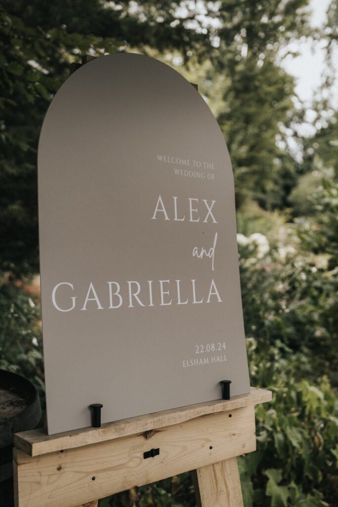 A taupe arched wedding sign on a wooden stand reads, “Welcome to the wedding of Alex and Gabriella, 22.08.24, Elsham Hall,” in white text. Captured by a wedding photographer, the sign sits outdoors amid lush greenery and soft natural light. © Aimee Lince Photography, Lincolnshire photographer