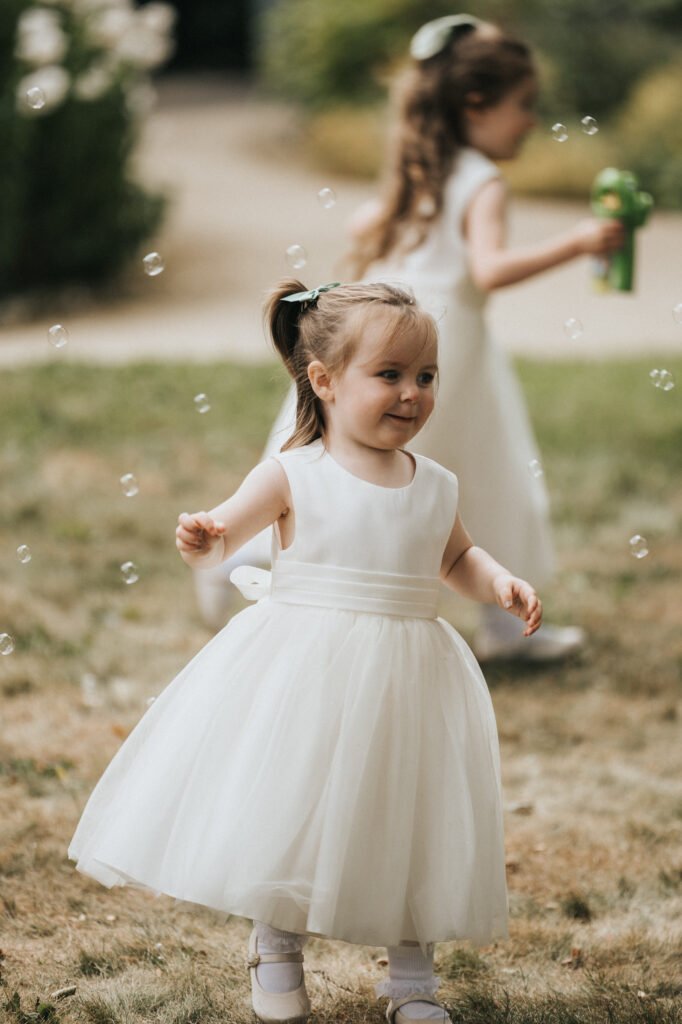 A young girl in a white dress smiles whilst playing outdoors with floating bubbles at Elsham Hall. Another girl, blurred in the background with a green bubble wand, captures a joyful moment perfect for a wedding photographer. © Aimee Lince Photography, Lincolnshire photographer