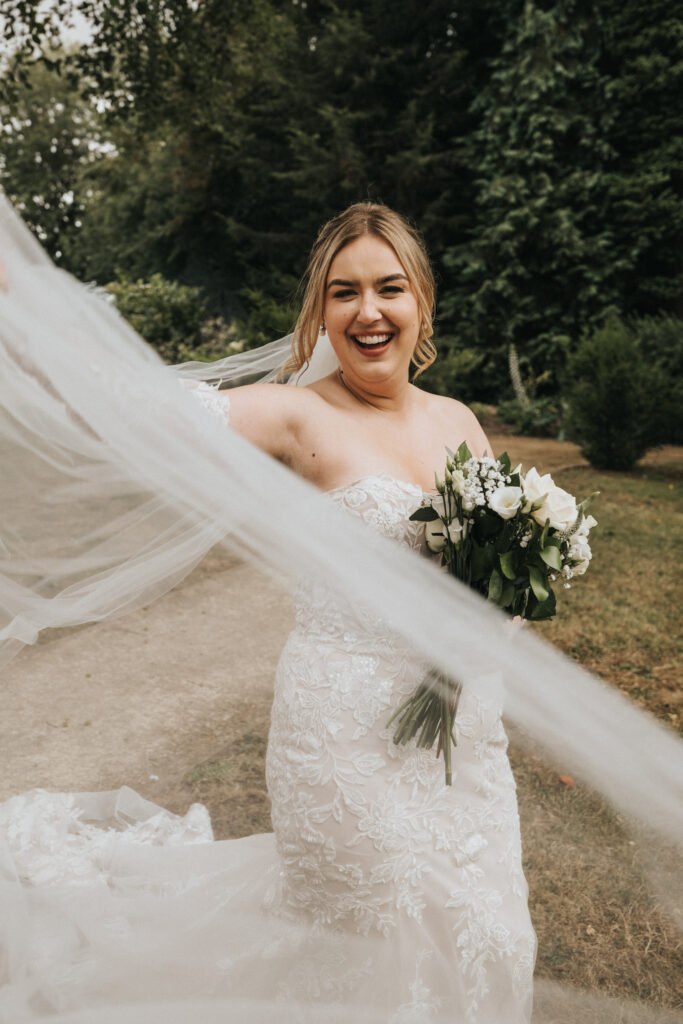 A joyful bride in a white lace dress holds a bouquet of white flowers and greenery. She smiles broadly outdoors at Elsham Hall, lifting her flowing veil, with trees and grass in the background—a candid moment captured by a wedding photographer. © Aimee Lince Photography, Lincolnshire photographer