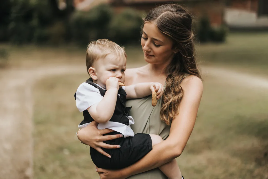 A young woman with long, wavy hair holds a small boy in a white shirt and dark waistcoat outside Elsham Hall. As the boy eats a biscuit, looking serious, the woman smiles gently—captured beautifully by their wedding photographer on the grassy grounds. © Aimee Lince Photography, Lincolnshire photographer