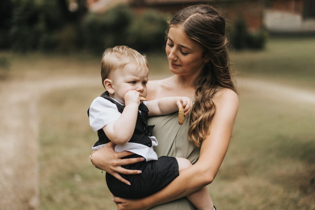 A young woman with long, wavy hair holds a small boy in a white shirt and dark waistcoat outside Elsham Hall. As the boy eats a biscuit, looking serious, the woman smiles gently—captured beautifully by their wedding photographer on the grassy grounds. © Aimee Lince Photography, Lincolnshire photographer
