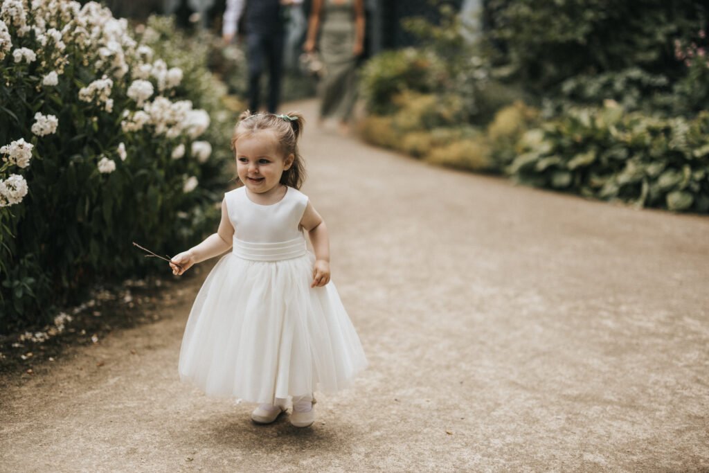 A young girl in a white, sleeveless, knee-length dress and white shoes walks happily along a garden path at Elsham Hall, lined with green bushes and white flowers. She holds a stick and smiles, with blurred adults—perhaps the wedding photographer—in the background. © Aimee Lince Photography, Lincolnshire photographer