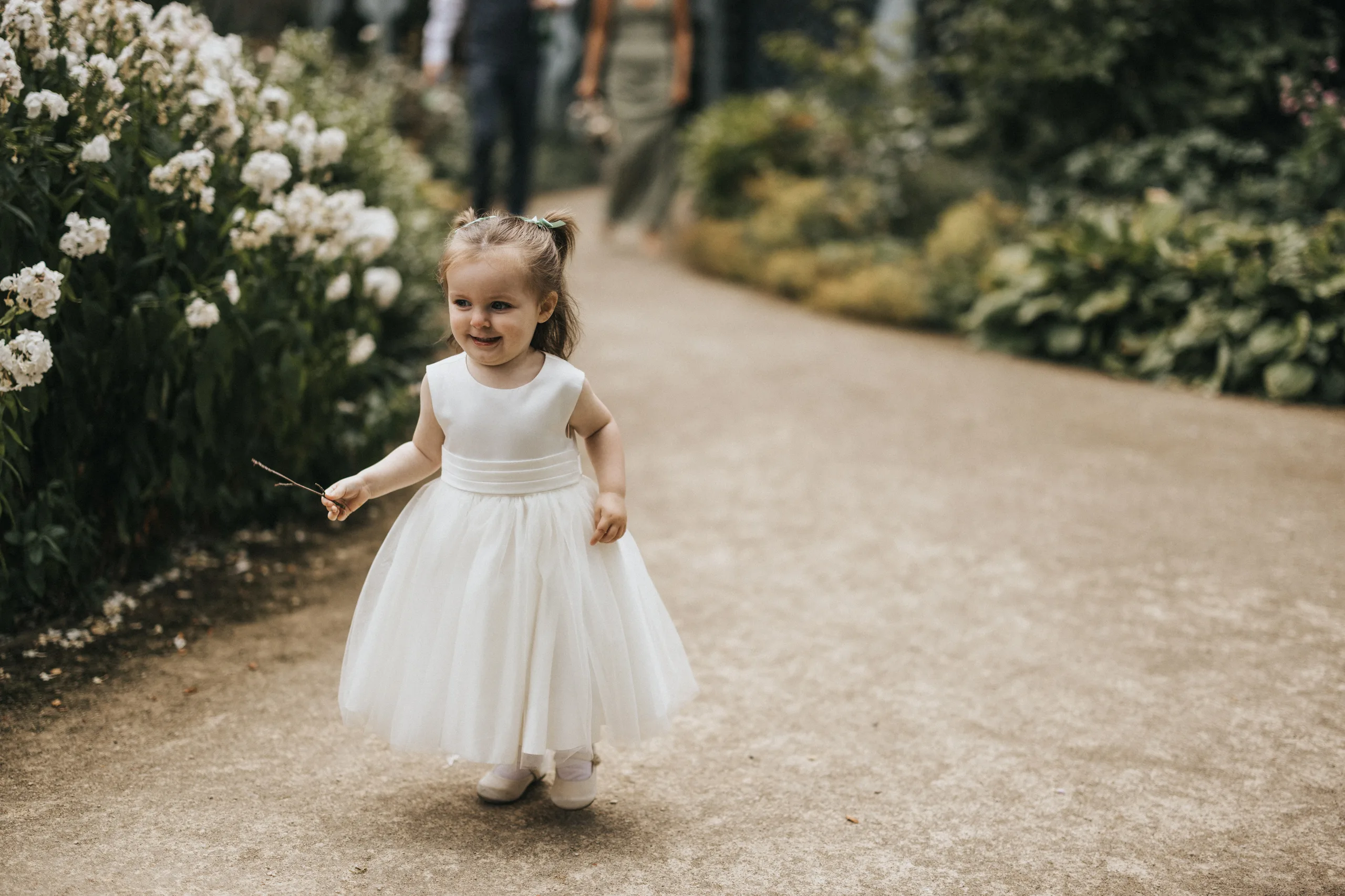 A young girl in a white, sleeveless, knee-length dress and white shoes walks happily along a garden path at Elsham Hall, lined with green bushes and white flowers. She holds a stick and smiles, with blurred adults—perhaps the wedding photographer—in the background. © Aimee Lince Photography, Lincolnshire photographer