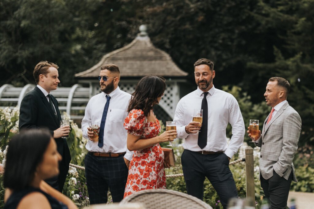 Five people dressed in formal attire stand outdoors at Elsham Hall, socialising and holding drinks. Captured by a wedding photographer, they mingle among lush greenery and a wooden gazebo in the background. © Aimee Lince Photography, Lincolnshire photographer