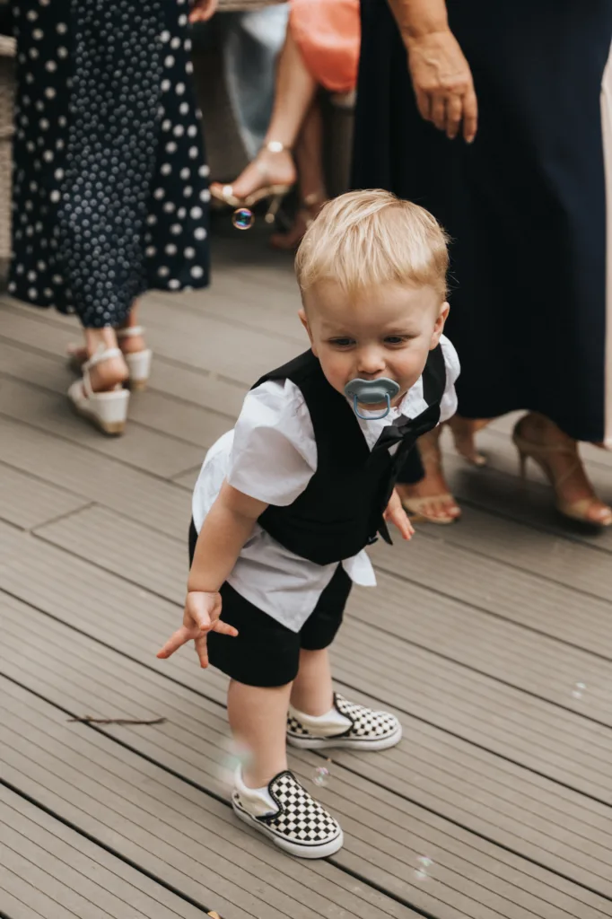 A toddler with blond hair, in a white shirt and black waistcoat, bends down on a wooden decking at Elsham Hall. A soap bubble floats near the child's hand as adults in dresses and heels look on—perfect for any wedding photographer to capture. © Aimee Lince Photography, Lincolnshire photographer