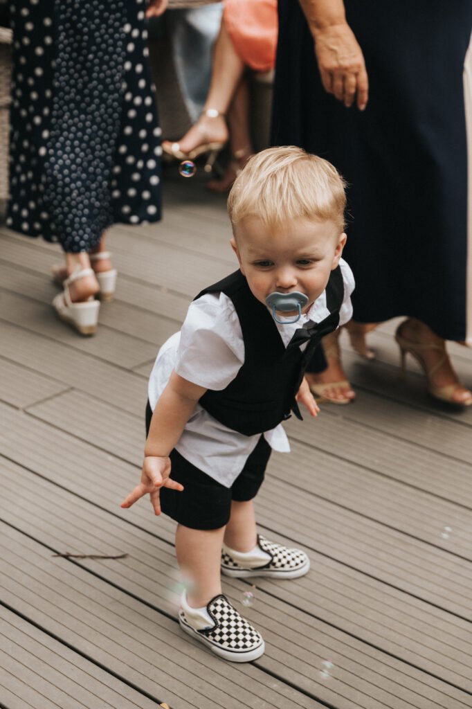 A toddler with blond hair, in a white shirt and black waistcoat, bends down on a wooden decking at Elsham Hall. A soap bubble floats near the child's hand as adults in dresses and heels look on—perfect for any wedding photographer to capture. © Aimee Lince Photography, Lincolnshire photographer