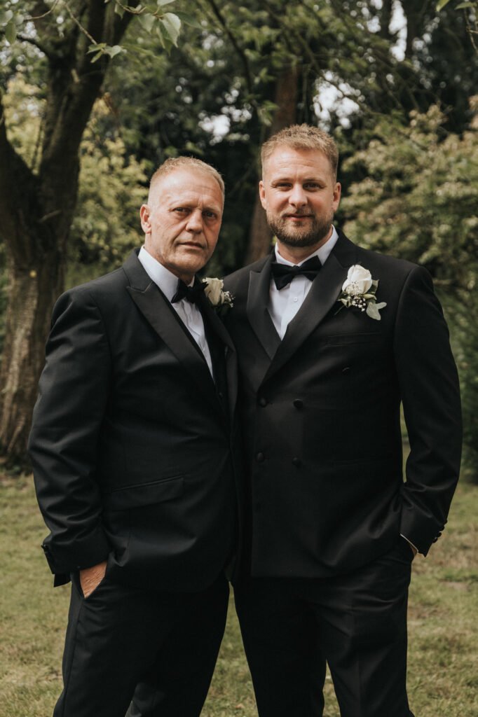 Two men in black dinner jackets with white buttonholes stand side by side outdoors at Elsham Hall, one older with grey hair and the other younger with a trimmed beard. Captured by a wedding photographer, they pose before leafy trees with serious expressions. © Aimee Lince Photography, Lincolnshire photographer