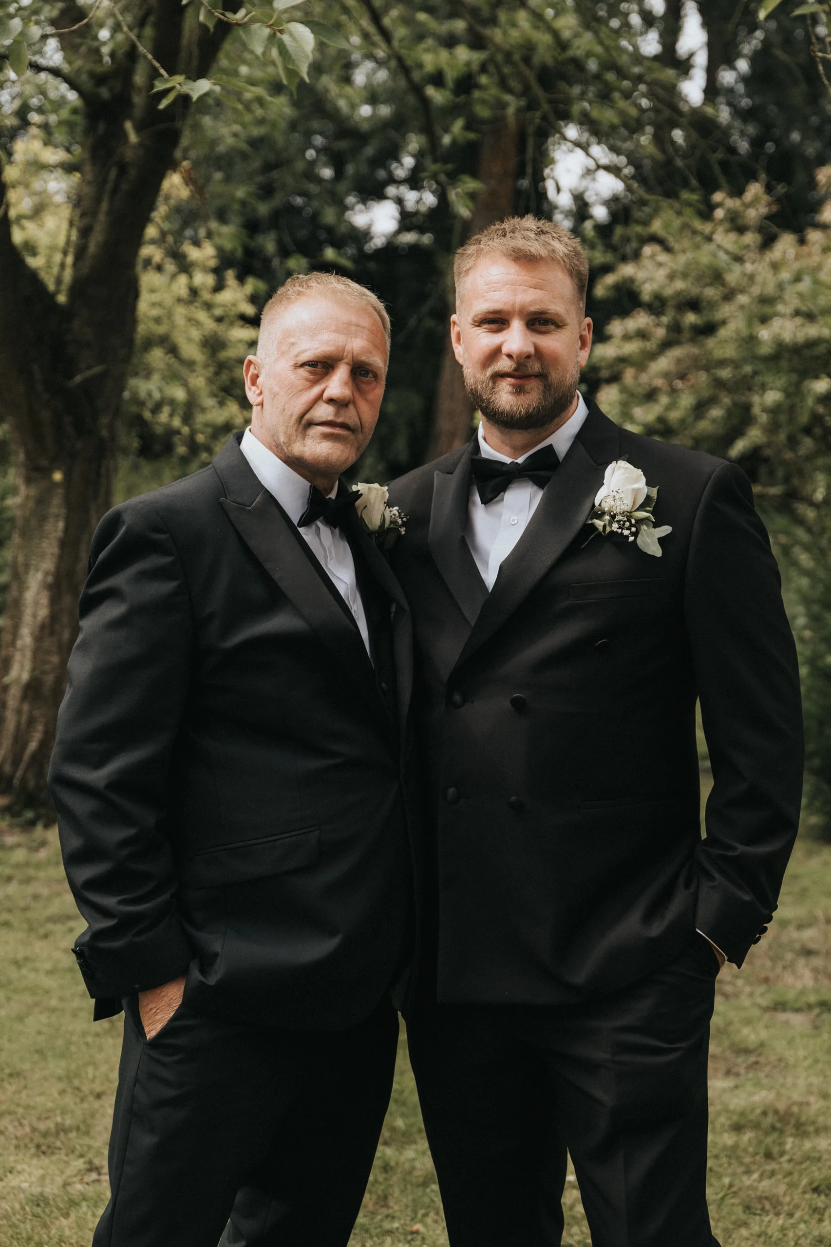 Two men in black dinner jackets with white buttonholes stand side by side outdoors at Elsham Hall, one older with grey hair and the other younger with a trimmed beard. Captured by a wedding photographer, they pose before leafy trees with serious expressions. © Aimee Lince Photography, Lincolnshire photographer