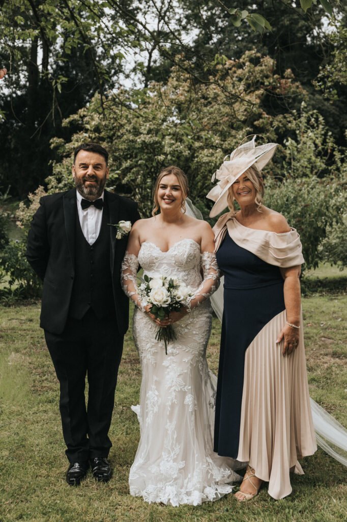 Three adults stand outdoors on grass, smiling. At the centre, a bride in a lace gown holds a bouquet, joined by loved ones. Captured by a wedding photographer at Elsham Hall, lush greenery forms the perfect backdrop for this joyful moment. © Aimee Lince Photography, Lincolnshire photographer