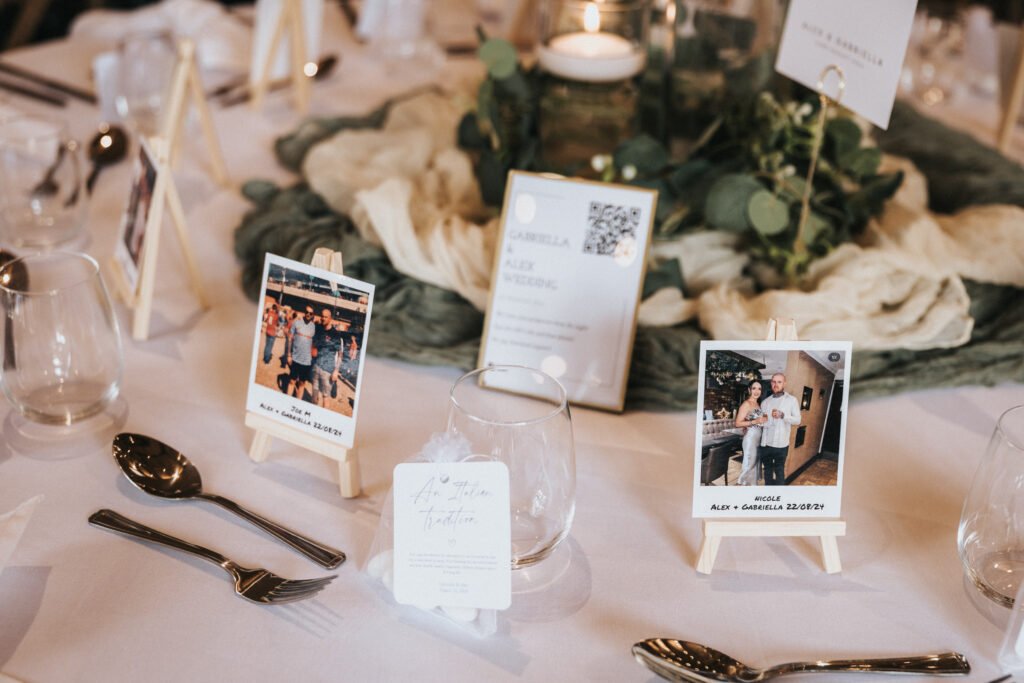 A decorated table at Elsham Hall set for an event features glassware, cutlery, candles, greenery, and small easels displaying Polaroid-style photos by the wedding photographer, name cards, and a QR code for a personalised and elegant atmosphere. © Aimee Lince Photography, Lincolnshire photographer