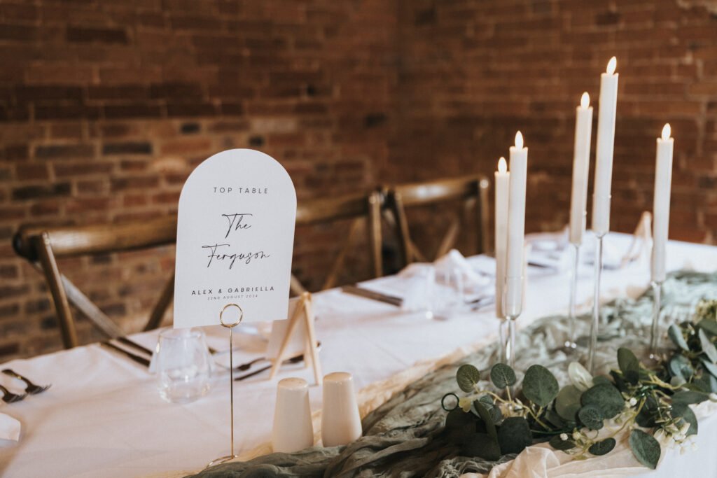 A wedding top table at Elsham Hall is decorated with tall white candles in glass holders, green foliage, and a sign reading "The Fergusons." Perfect for a wedding photographer, the setting features glassware and serviettes against a rustic brick wall. © Aimee Lince Photography, Lincolnshire photographer
