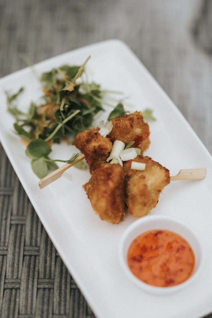 A close-up of breaded, deep-fried skewered pieces on a white plate at Elsham Hall, garnished with chopped spring onions, served with orange dipping sauce and fresh mixed salad—perfect for a wedding photographer capturing delicious moments. © Aimee Lince Photography, Lincolnshire photographer