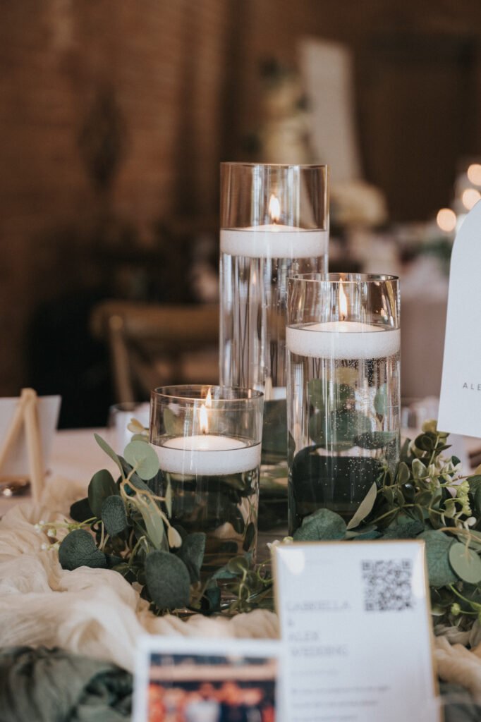 Three glass vases of varying heights, each with a floating white candle, sit on a table adorned with greenery and soft white fabric—an elegant setup perfect for an Elsham Hall celebration, beautifully captured by a wedding photographer. © Aimee Lince Photography, Lincolnshire photographer