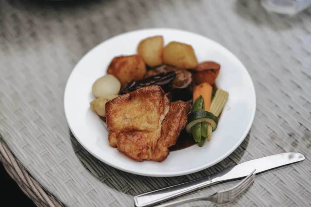 A white plate holds a roast dinner with Yorkshire pudding, roast potatoes, and meat with dark gravy—an inviting scene at Elsham Hall, perfect for a wedding photographer to capture. The plate rests on a woven table with a knife and fork beside it. © Aimee Lince Photography, Lincolnshire photographer