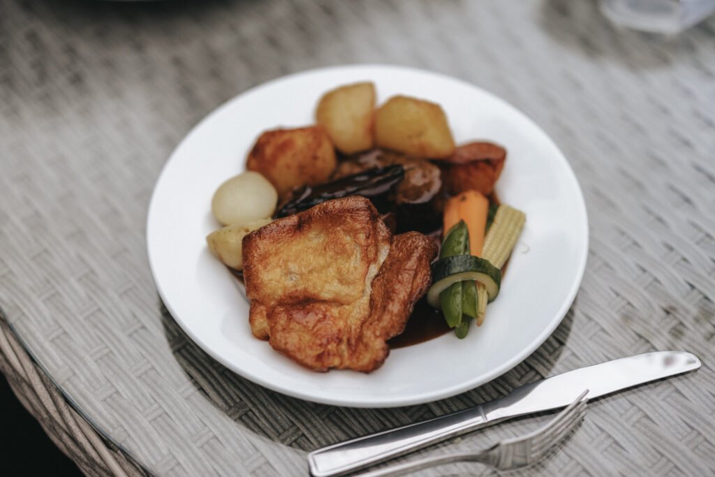 A white plate holds a roast dinner with Yorkshire pudding, roast potatoes, and meat with dark gravy—an inviting scene at Elsham Hall, perfect for a wedding photographer to capture. The plate rests on a woven table with a knife and fork beside it. © Aimee Lince Photography, Lincolnshire photographer