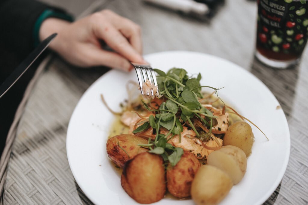 A hand holds a fork over a white plate with roast potatoes, boiled potatoes, salmon topped with green herbs, and leafy greens—an elegant meal perfect for a wedding photographer capturing special moments at Elsham Hall. © Aimee Lince Photography, Lincolnshire photographer