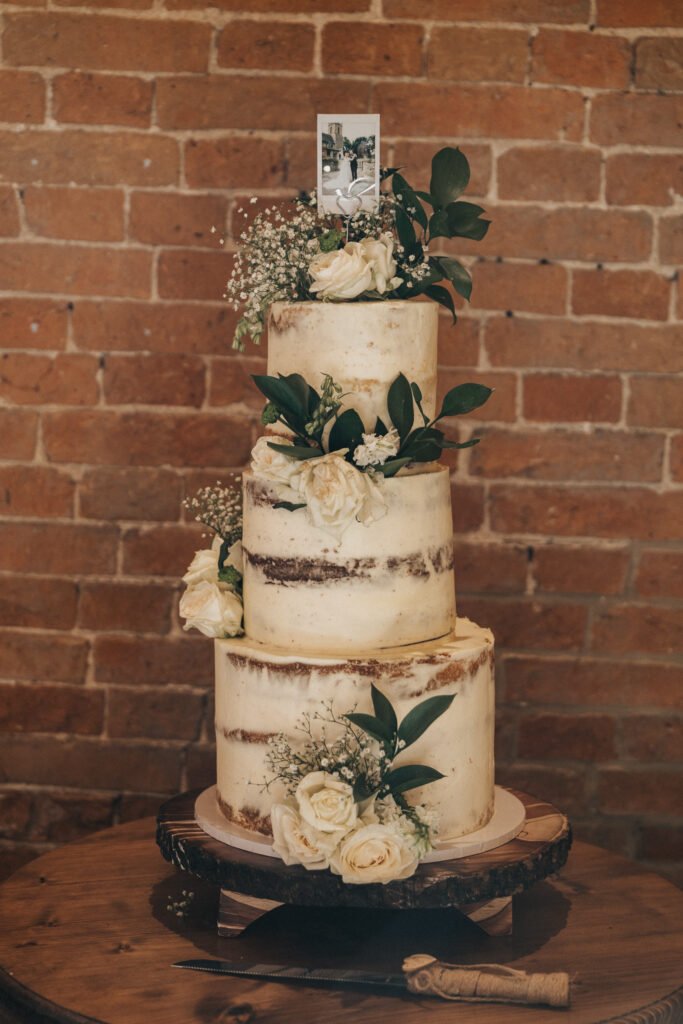 A three-tiered semi-naked wedding cake decorated with white roses, greenery, and baby's breath sits on a wooden stand in front of a rustic brick wall at Elsham Hall. A wedding photographer captures the scene, complete with photo topper and cake knife nearby. © Aimee Lince Photography, Lincolnshire photographer