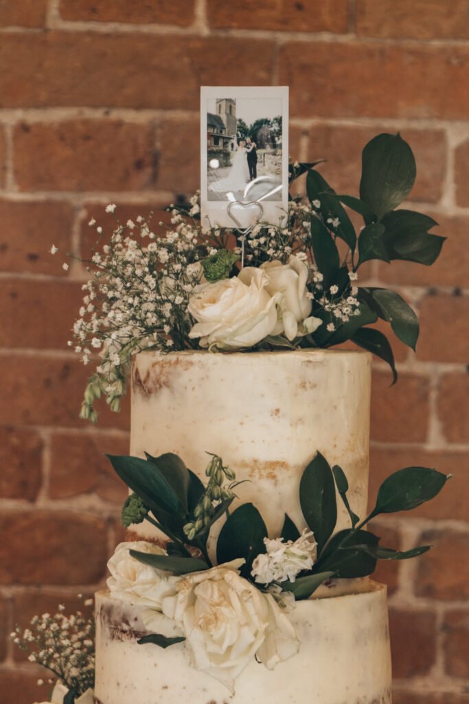 A semi-naked two-tier wedding cake adorned with white roses, greenery, and baby's breath, topped with a couple's photo on a metal holder. Perfect for an Elsham Hall celebration, it sits beautifully against a rustic exposed brick wall—ideal for any wedding photographer. © Aimee Lince Photography, Lincolnshire photographer