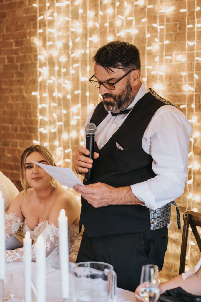 A man in a black waistcoat and glasses reads from a paper into a microphone at a table, with a woman in a white off-the-shoulder dress beside him. Warm fairy lights and candles set the mood—an intimate moment beautifully captured by the wedding photographer at Elsham Hall. © Aimee Lince Photography, Lincolnshire photographer