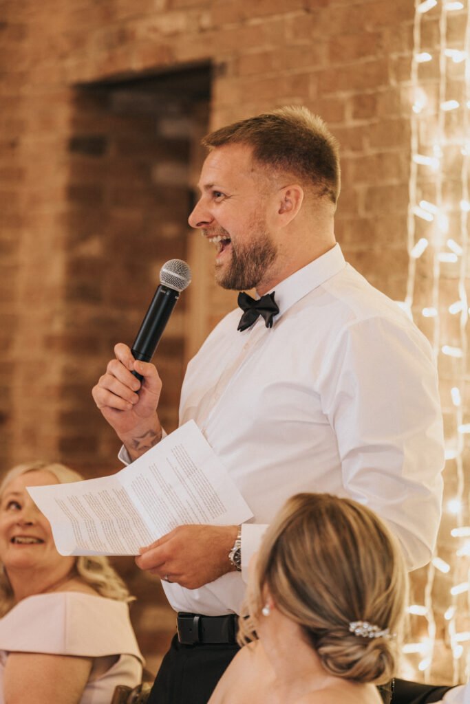 A wedding photographer at Elsham Hall captures a man in a white shirt and black bow tie, smiling as he gives a speech with microphone and papers, whilst two women look up at him. The brick wall and fairy lights create a warm, festive ambience. © Aimee Lince Photography, Lincolnshire photographer