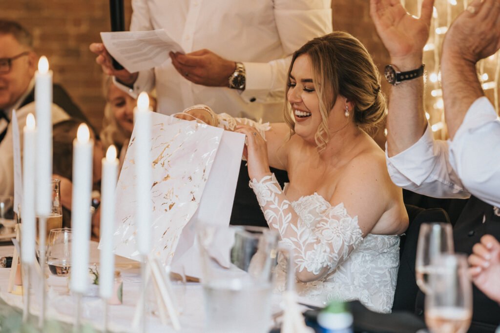 A smiling bride in a white lace off-shoulder gown sits at a table, opening a large gift bag. Captured by a wedding photographer at Elsham Hall, she’s surrounded by guests and candles in a joyful, warmly lit celebration. © Aimee Lince Photography, Lincolnshire photographer