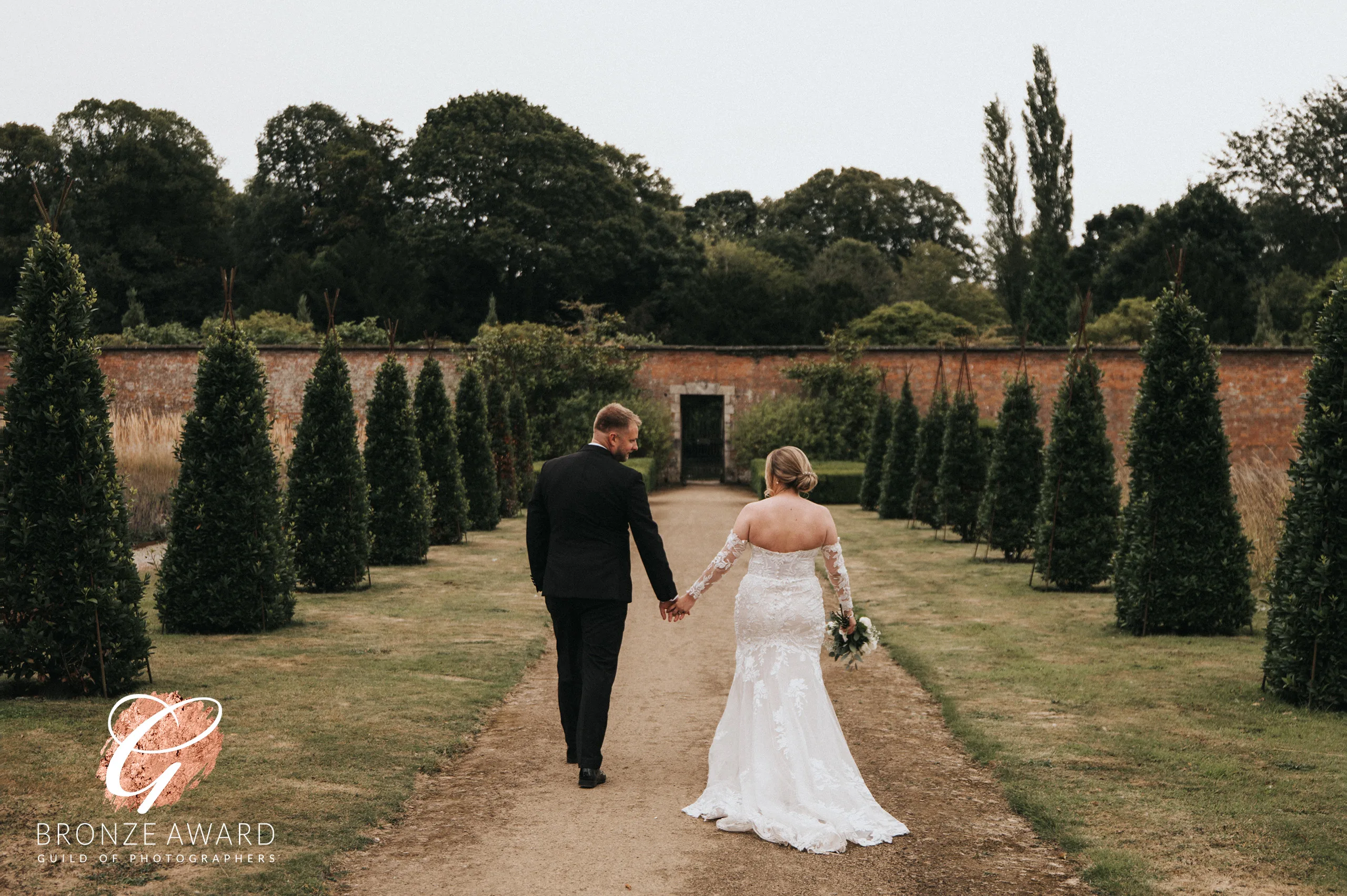 A bride in a white lace gown and a groom in a black suit walk hand-in-hand down a garden path at Elsham Hall, captured by a wedding photographer. The bride holds a small bouquet, with trimmed trees, greenery, and a brick wall in the background. © Aimee Lince Photography, Lincolnshire photographer