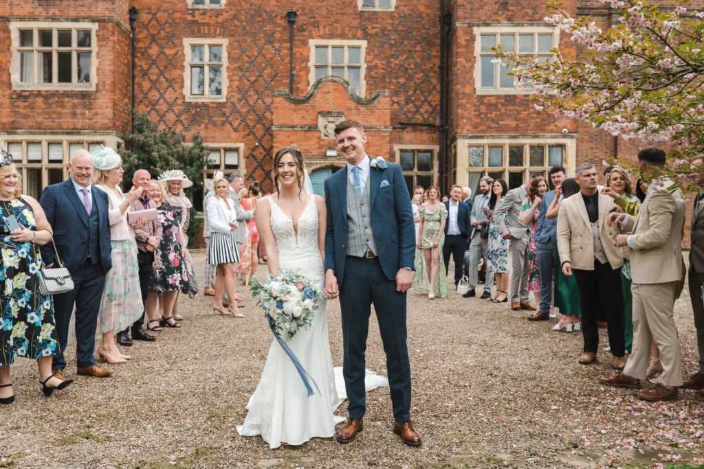 A bride and groom stand smiling in front of a historic brick building, surrounded by guests in colourful outfits, with blooming trees and spring flowers in the background. © Aimee Lince Photography, wedding and lifestyle photographer in Lincolnshire