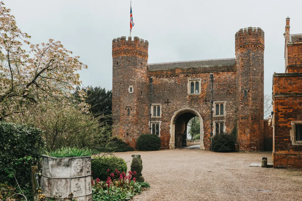 An historic brick gatehouse with two round towers, arched entryway, and a British flag on top, surrounded by greenery and gardens on a cloudy day. © Aimee Lince Photography, wedding and lifestyle photographer in Lincolnshire