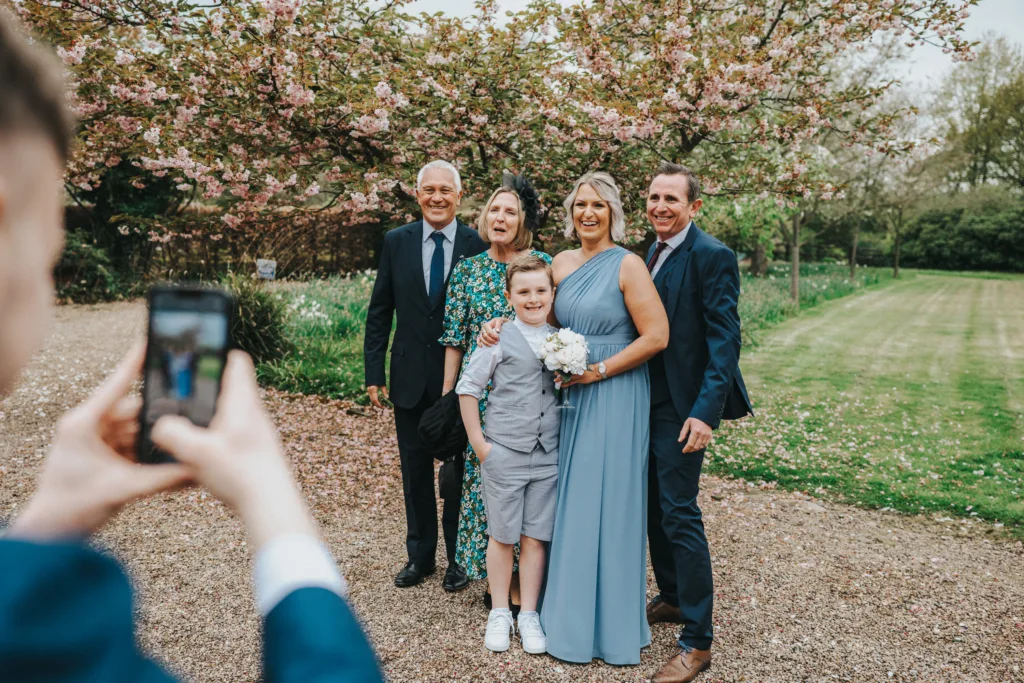 A group of five people dressed formally pose and smile for a photo outdoors under blossoming trees, while a child in the foreground takes their picture with a mobile phone. © Aimee Lince Photography, wedding and lifestyle photographer in Lincolnshire