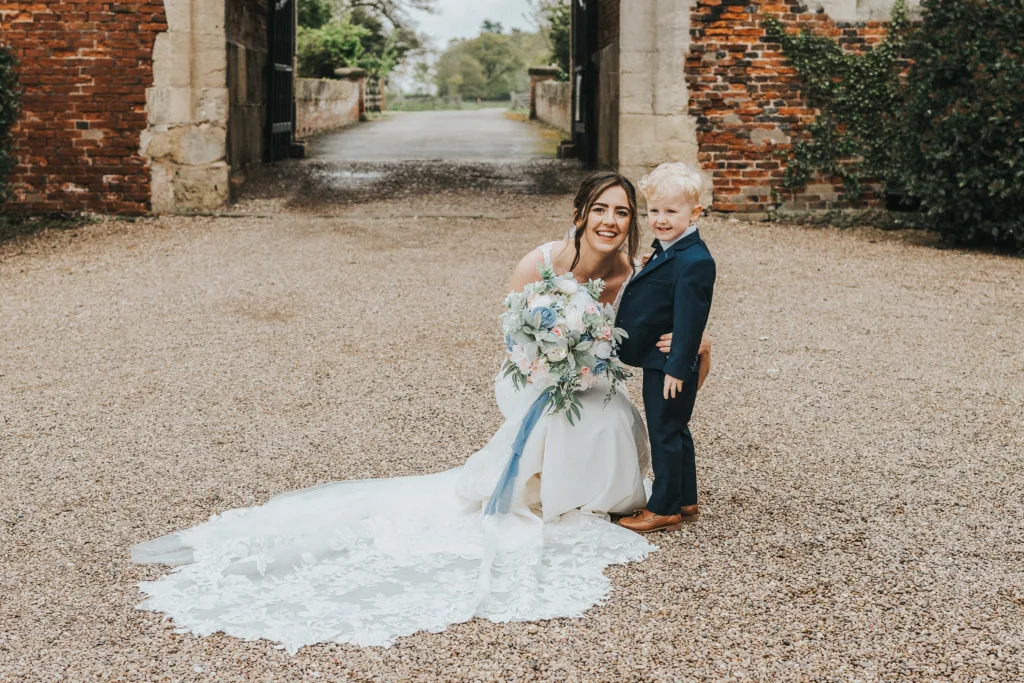 A smiling bride in a white gown and holding a bouquet kneels beside a young boy in a navy suit, posing together outdoors on a gravel path with brick walls and greenery in the background. © Aimee Lince Photography, wedding and lifestyle photographer in Lincolnshire