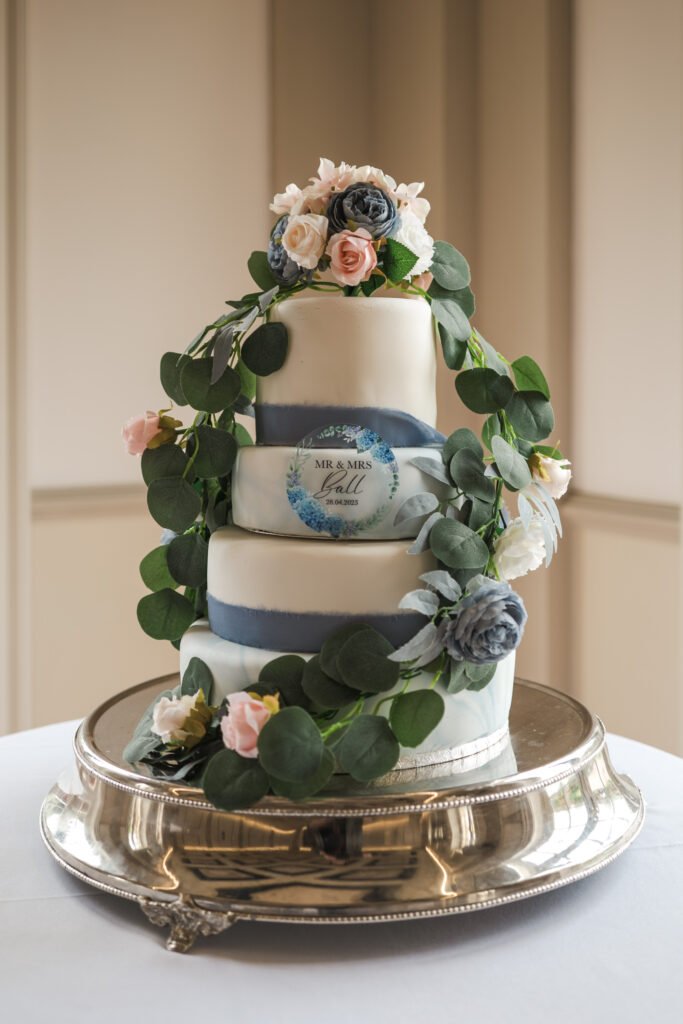 A three-tiered wedding cake decorated with pink and grey flowers, greenery, and blue ribbons, sits on a silver platter. The middle tier features a sign reading “MR & MRS Hall.”. © Aimee Lince Photography, wedding and lifestyle photographer in Lincolnshire
