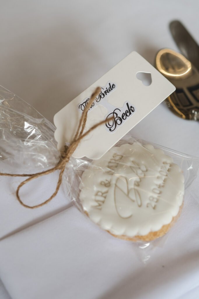 A white iced biscuit with "MR & MRS Beck" and a date embossed on it, wrapped in clear plastic with twine and a tag that reads "The Bride Beck," placed on a white tablecloth near gold cutlery. © Aimee Lince Photography, wedding and lifestyle photographer in Lincolnshire