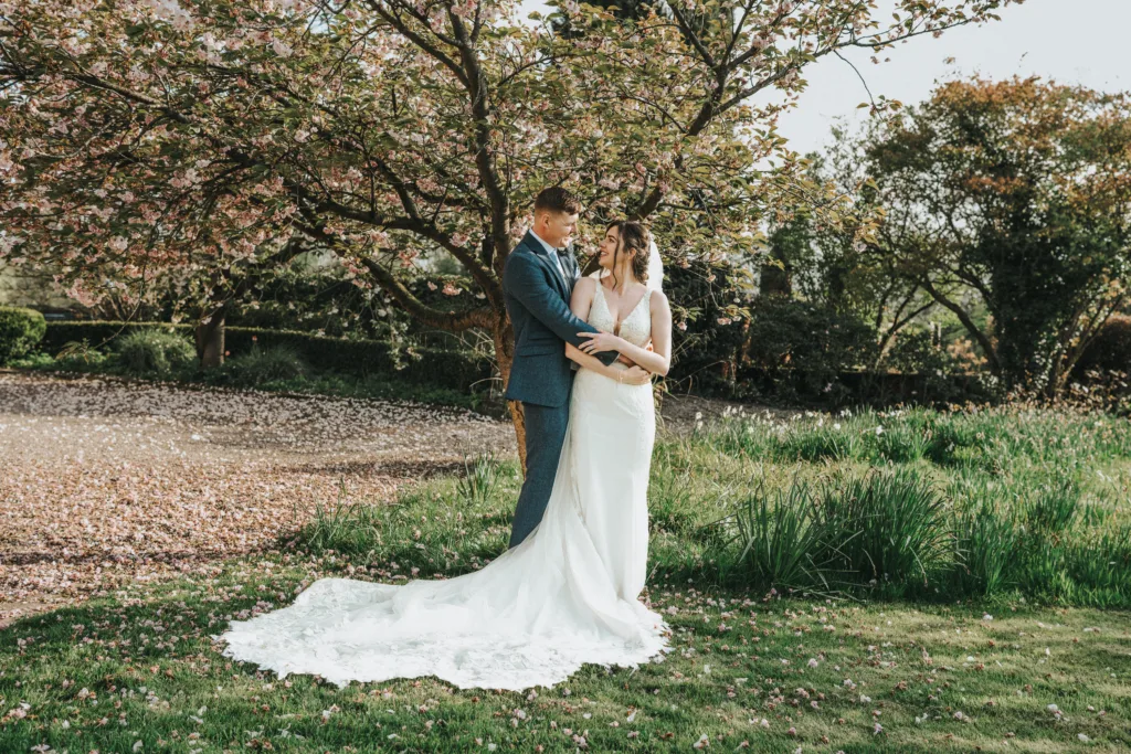 A bride and groom stand together outdoors under a blossoming tree, embracing and smiling at each other. The bride wears a long white dress, and the groom is in a blue suit. Sunlight filters through the trees around them. © Aimee Lince Photography, wedding and lifestyle photographer in Lincolnshire