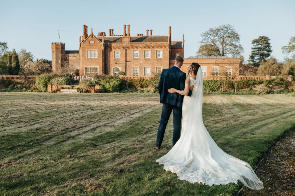 A bride in a long white dress and veil stands arm in arm with a groom in a dark suit, both facing a large historic mansion surrounded by gardens and trees on a sunny day. © Aimee Lince Photography, wedding and lifestyle photographer in Lincolnshire
